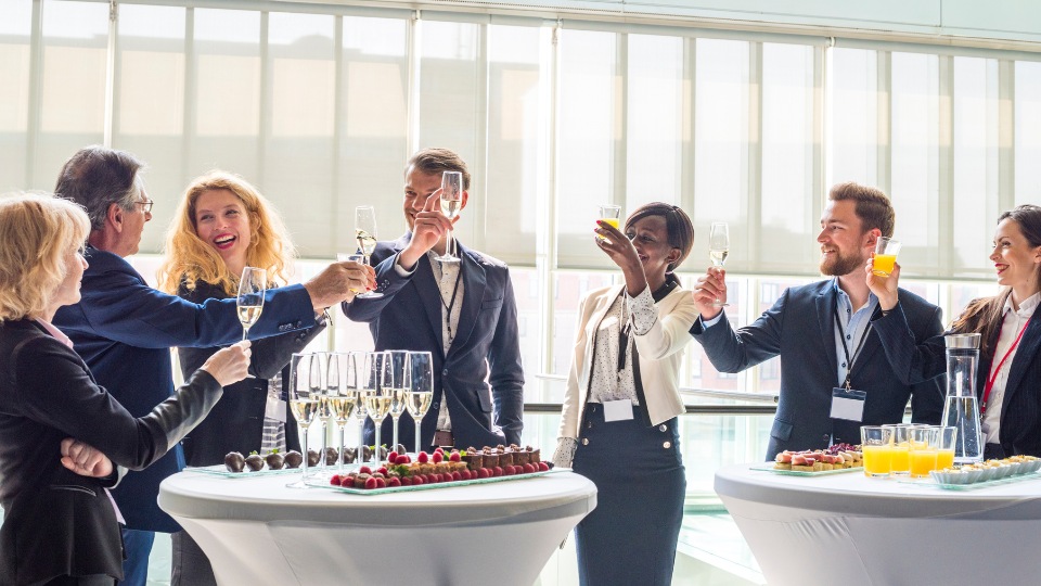 Un grupo de seis personas diversas vestidas formalmente brindan con bebidas alrededor de mesas llenas de bocadillos y bebidas. Ambiente luminoso y alegre.