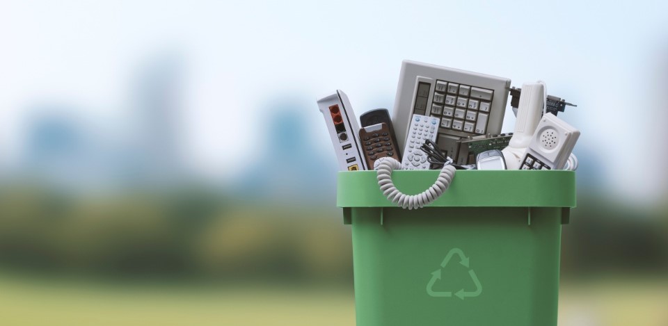 En primer plano una basura verde con el símbolo de reciclaje llena de aparatos y dispositivos electrónicos. 