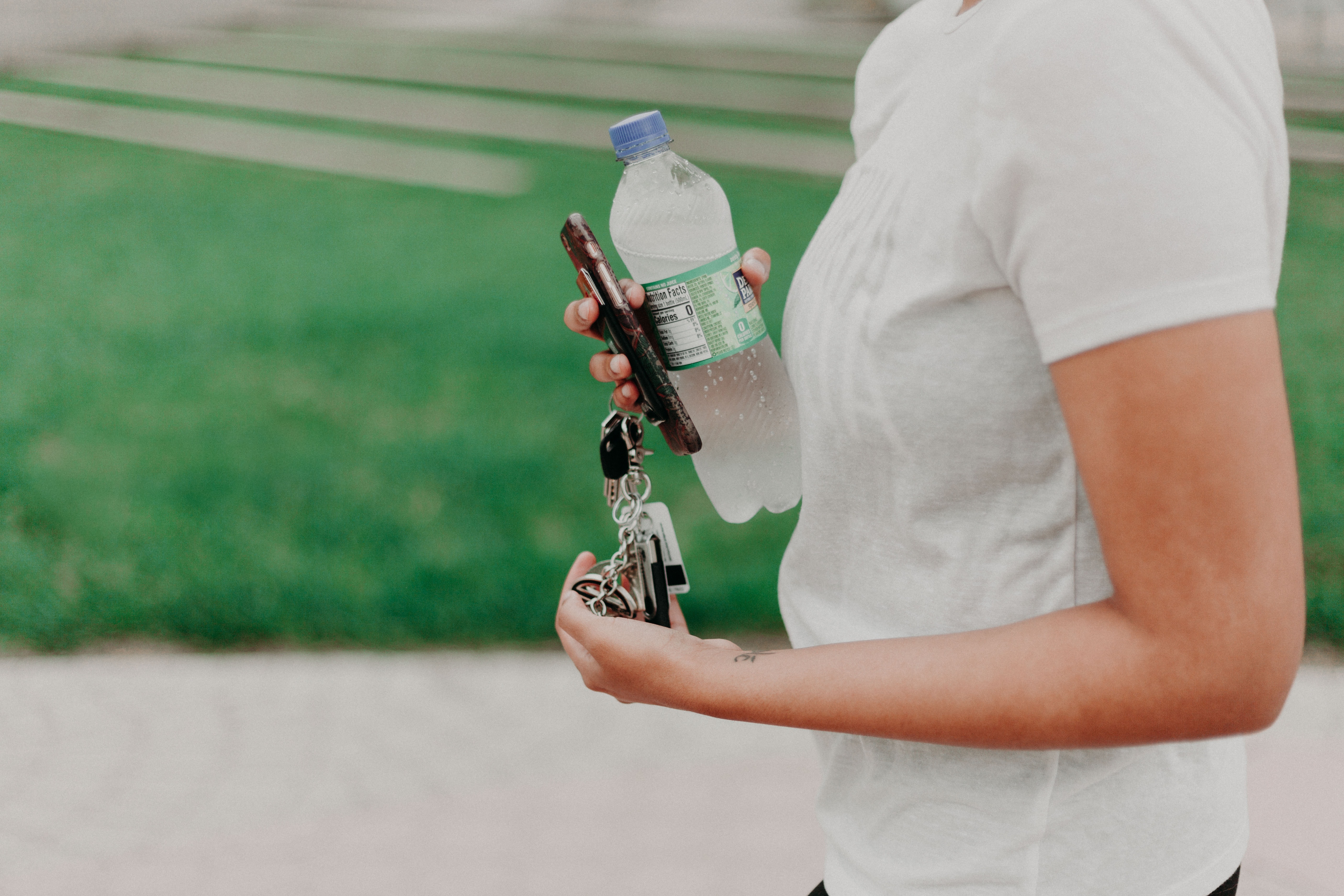 Imagen de la parte del cuerpo de una mujer con una botella de agua, llaves y un móvil en las manos, detrás un campo verde difuminado 