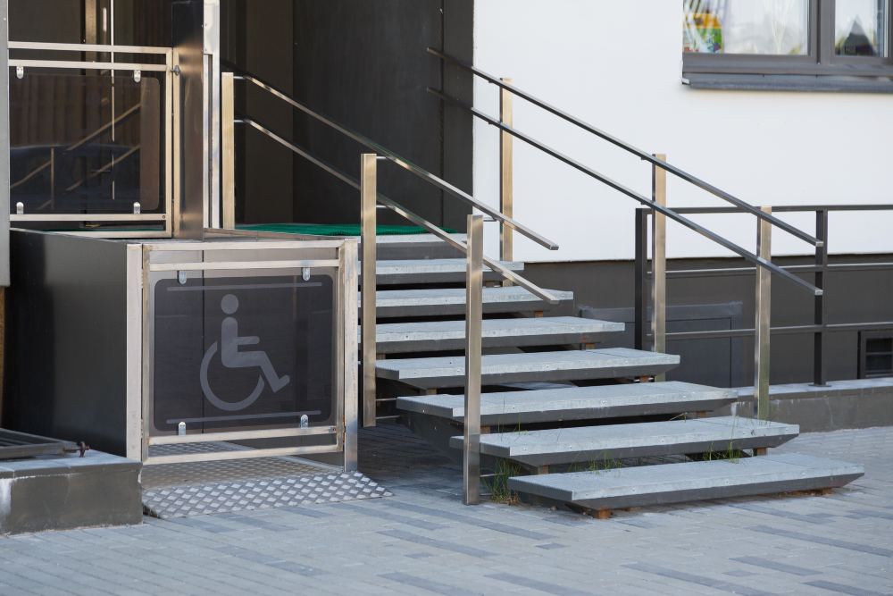 Entrada a un edificio con rampa accesible y escaleras, barandillas metálicas y señal de acceso para personas en silla de ruedas.