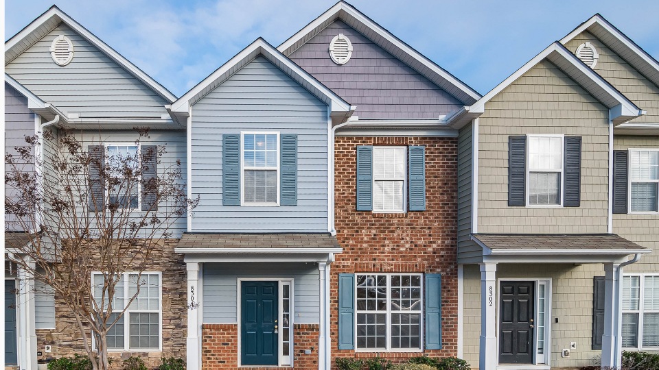 Hilera de tres casas adosadas bajo un cielo azul, cada una con fachadas diferentes: revestimiento gris, ladrillo rojo y revestimiento beige, creando una sensación de vecindario armonioso.