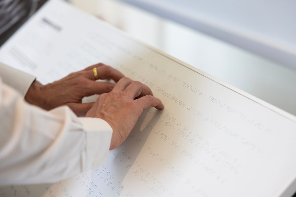 Primer plano de unas manos leyendo un documento en braille sobre una superficie blanca. Los dedos recorren las líneas de puntos en relieve para interpretar el contenido táctil. La imagen pone el foco en la accesibilidad de la información para personas con discapacidad visual.