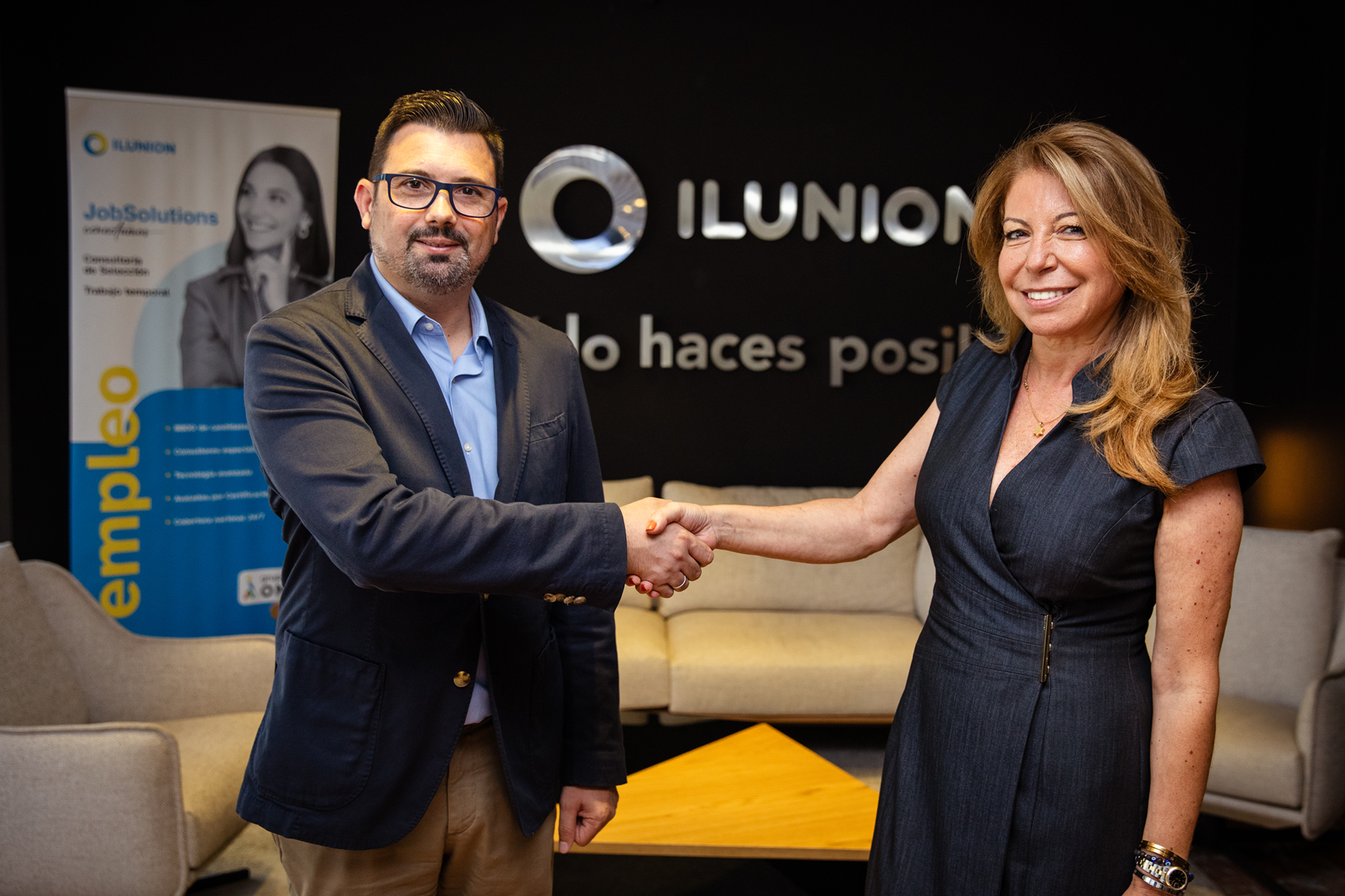 Dos profesionales estrechan la mano frente al logotipo de ILUNION y el lema “Tú o haces posible”, con un roll‑up de empleo al fondo.