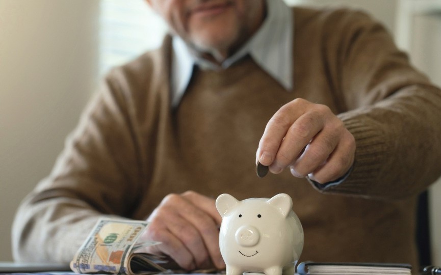 Una persona mayor con un suéter marrón coloca una moneda en una alcancía blanca, lo que da la impresión de estar ahorrando. Cerca de la mesa hay un fajo de billetes de dólar.