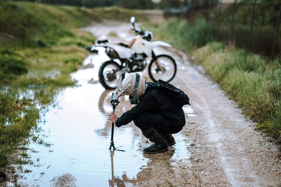 Persona con casco usando un trípode en un camino de tierra con charcos, con una motocicleta blanca al fondo.