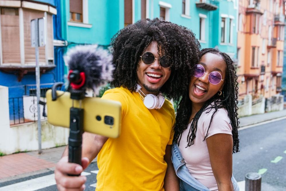 Dos personas posan para un selfie con un móvil en un estabilizador en una calle con edificios coloridos.