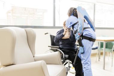 Una profesional de ILUNION VidaSénior se dispone a levantar a una mujer mayor de su silla de ruedas para ayudarla a sentarse en un sillón