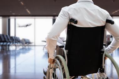 Fotografía de un hombre desplazándose en silla de ruedas por un aeropuerto. Al fondo, un avión despega.