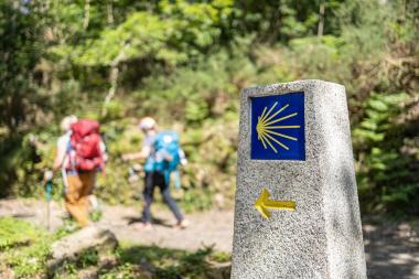 Dos personas al fondo realizan el camino de Santiago, indicado con una flecha y una concha sobre una piedra 