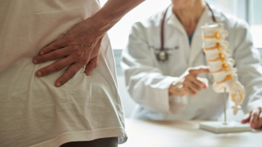 Persona con dolor lumbar en una consulta médica mientras un profesional sanitario muestra un modelo de columna vertebral.