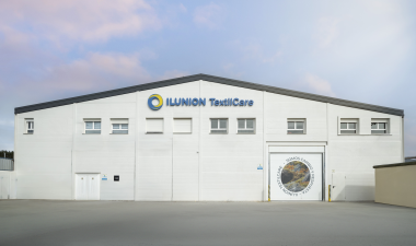 Exterior de una planta de ILUNION TextilCare. El edificio, de fachada blanca, muestra el logotipo de la empresa en la parte superior y varias ventanas. A la derecha hay una gran puerta de acceso con una imagen de paisaje y el mensaje “Somos cambio y respuesta”.