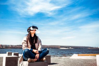 Una mujer joven en una playa con unas gafas de realidad virtual mirando hacia el cielo 