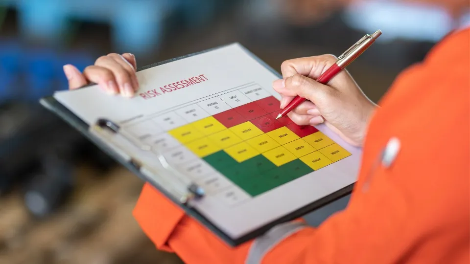 Una persona con una chaqueta naranja marca una sección en una tabla de evaluación de riesgos con código de colores en un portapapeles. La tabla incluye áreas rojas, amarillas y verdes.