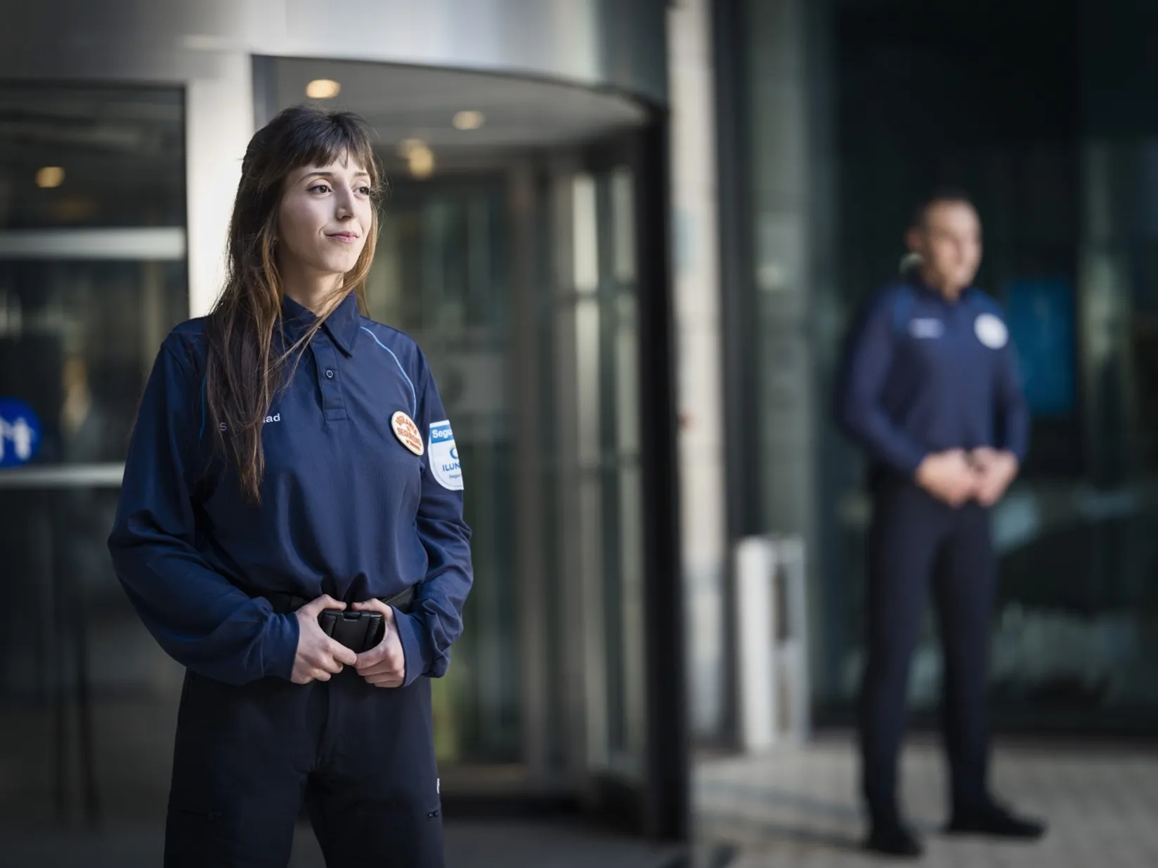 Dos vigilantes de seguridad de ILUNION, una mujer, en primer plano, y un hombre, custodian la entrada de un edificio