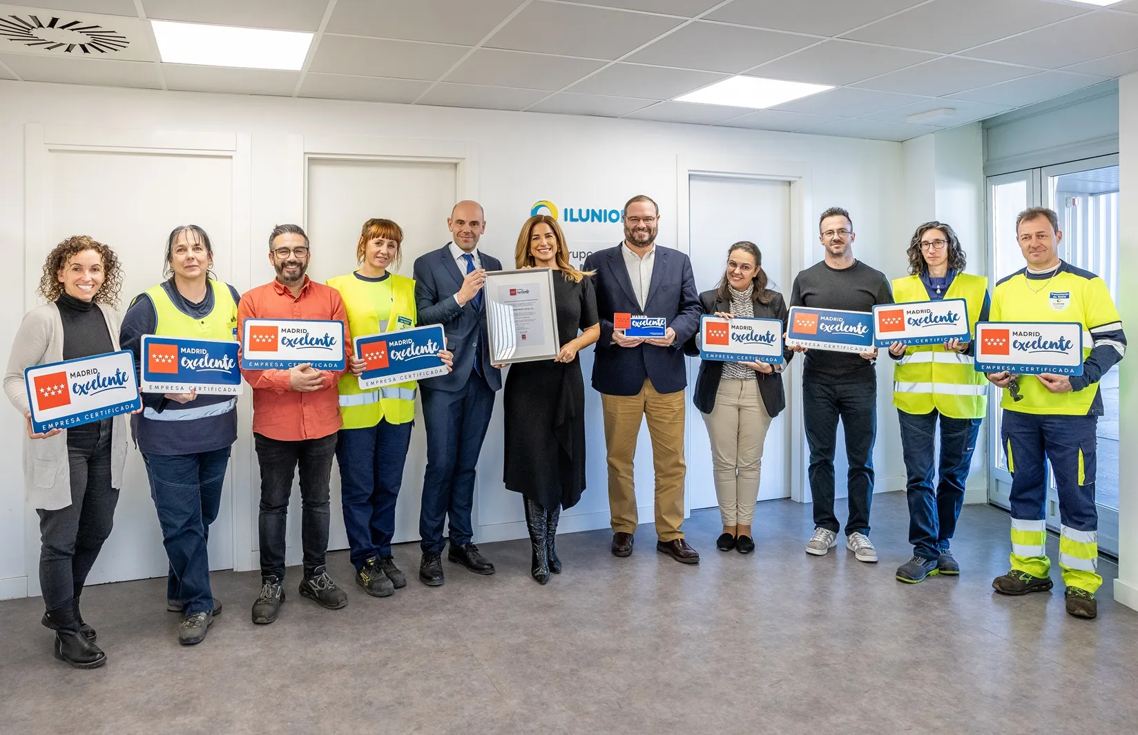 Un grupo de trabajadores de ILUNION Economía Circular, junto su director,  Pedro Antonio Martín, y la directora general de Madrid Excelente, Elena Mantilla. Ella sostiene la certificación de Madrid Excelente, y el resto de las personas tiene en sus manos un cartel donde se lee Madrid Excelente.   