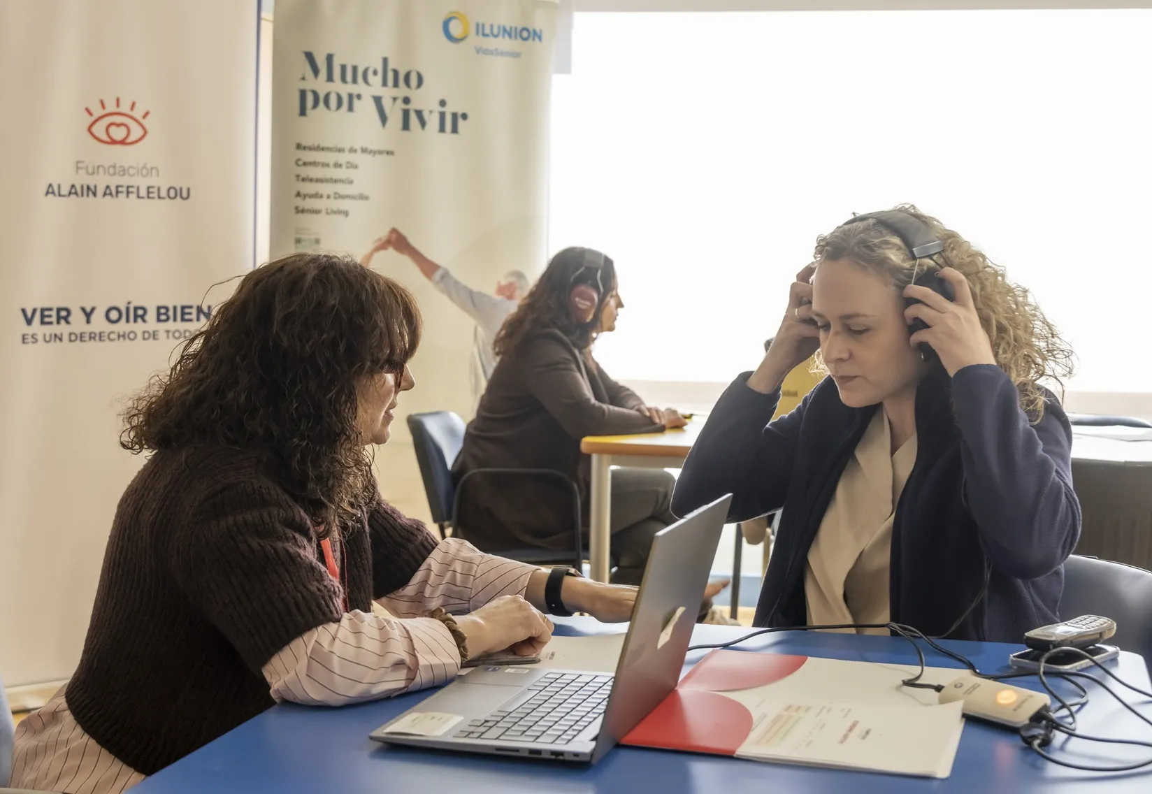 Una trabajadora de ILUNION VidaSénior se coloca los auriculares para realizarse una audiometría con una voluntaria de la Fundación Alain Afflelou