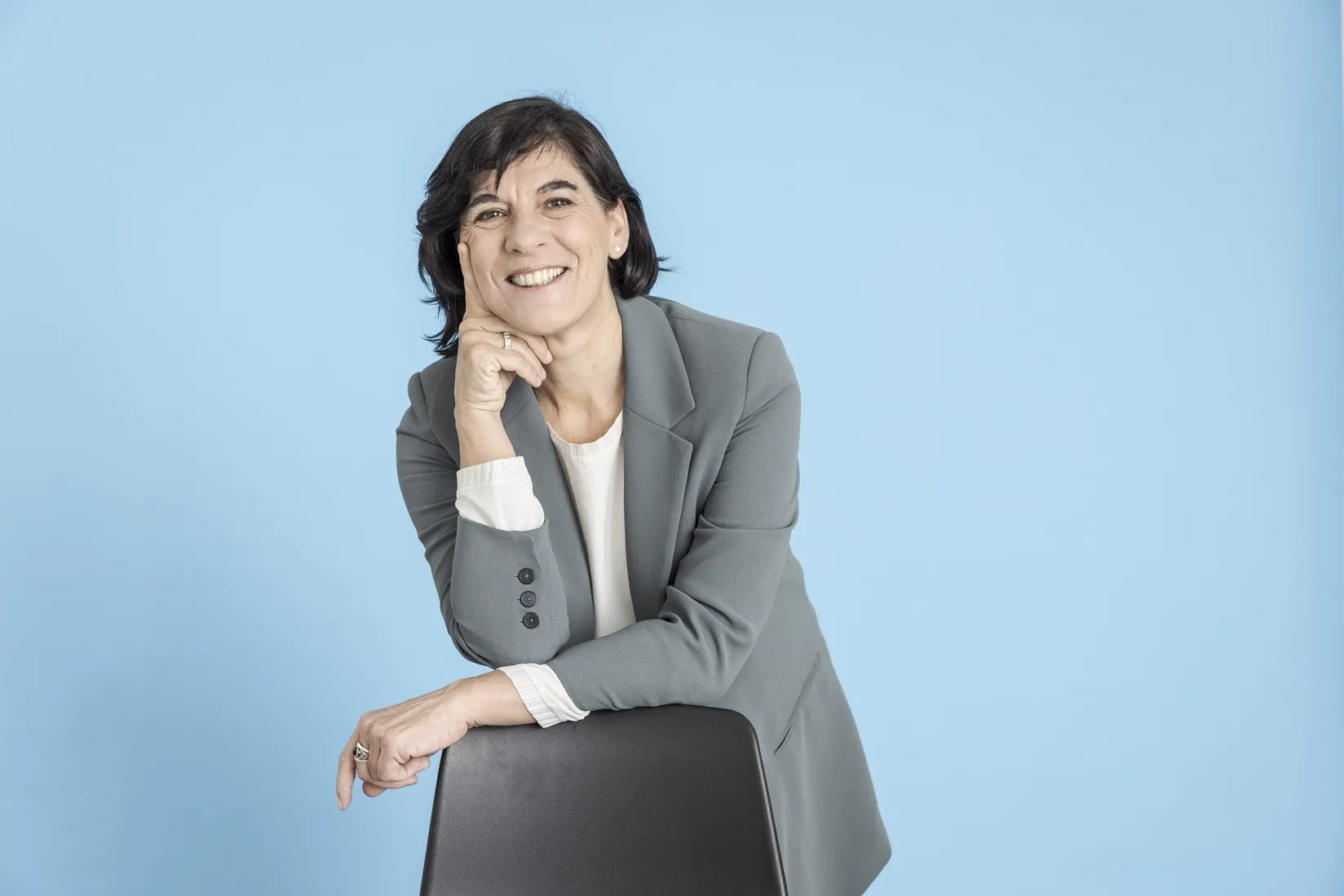 La imagen muestra un retrato de Pilar Soret en estudio con un fondo liso de color azul claro que ocupa toda la superficie posterior, generando un ambiente limpio y uniforme. En el primer plano aparece Pilar Soret apoyada ligeramente sobre el respaldo de una silla de color oscuro. El cuerpo está inclinado hacia delante, con el brazo izquierdo descansando sobre la silla y la mano derecha sosteniendo suavemente el mentón, en una postura relajada y natural.
