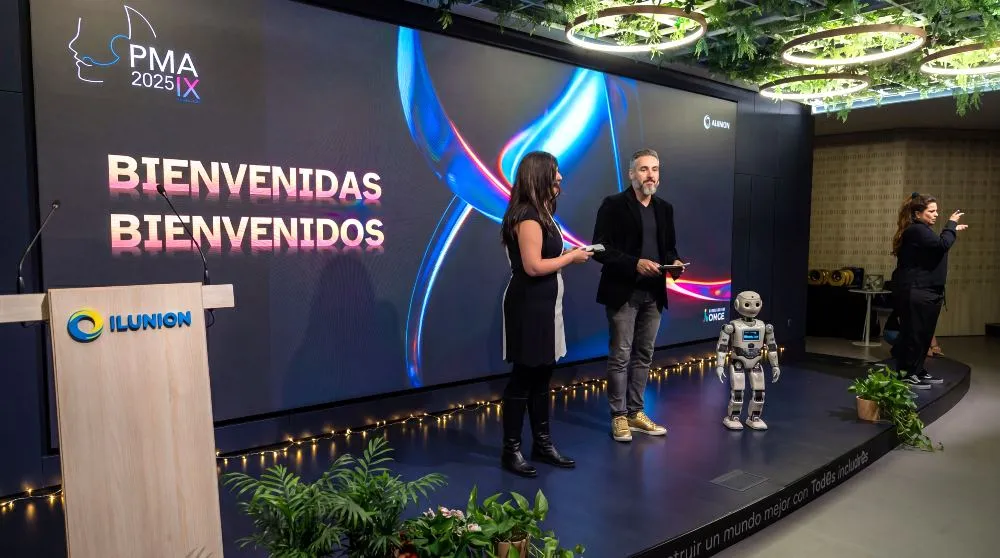 Escenario del evento PMA 2025 IX con el mensaje ‘Bienvenidas Bienvenidos’, dos ponentes y un robot humanoide junto al atril de ILUNION
