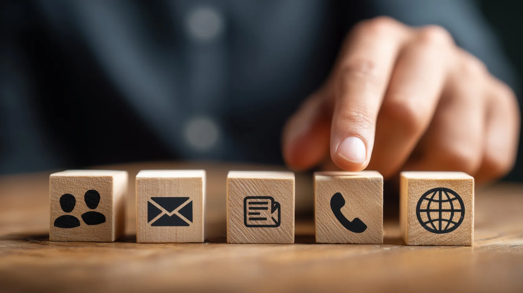 Primer plano de una mano seleccionando un bloque de madera con el icono de teléfono entre varios cubos con símbolos de canales de comunicación digital: usuarios, email, vídeo y web, sobre una superficie de madera.
