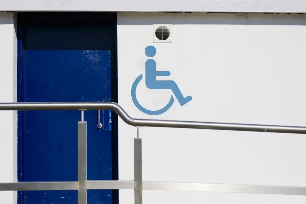 Símbolo azul de accesibilidad para silla de ruedas en pared blanca, junto a una puerta azul y barandilla metálica
