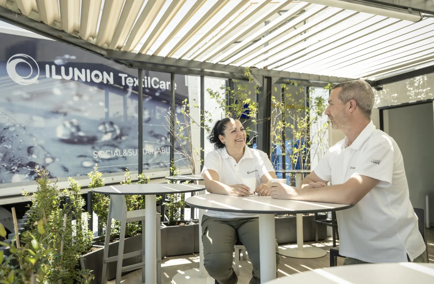 Dos personas que trabajan en ILUNION TextilCare charlan en una zona con sol y plantas de la compañía