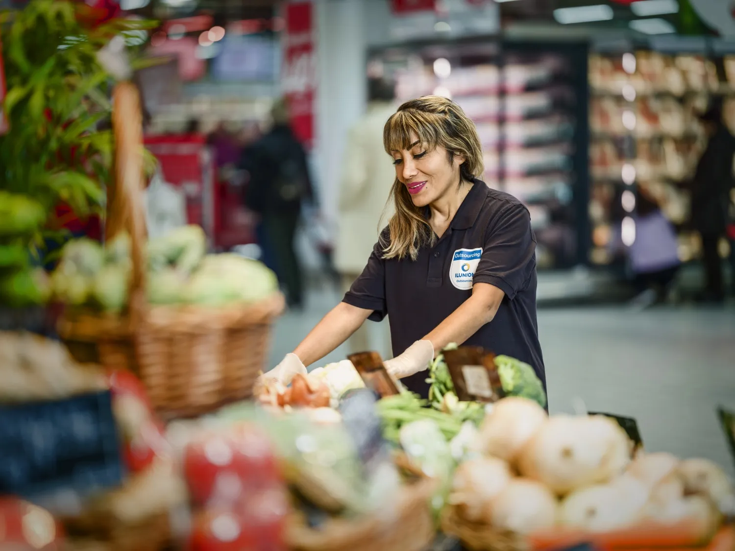 Empleado con guantes ordena verduras en un supermercado, con cestas de productos frescos desenfocadas en primer plano