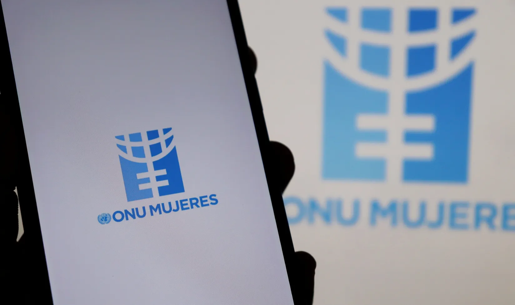Logotipo de ONU Mujeres en pantalla de un móvil, con versión ampliada y desenfocada del mismo fondo.