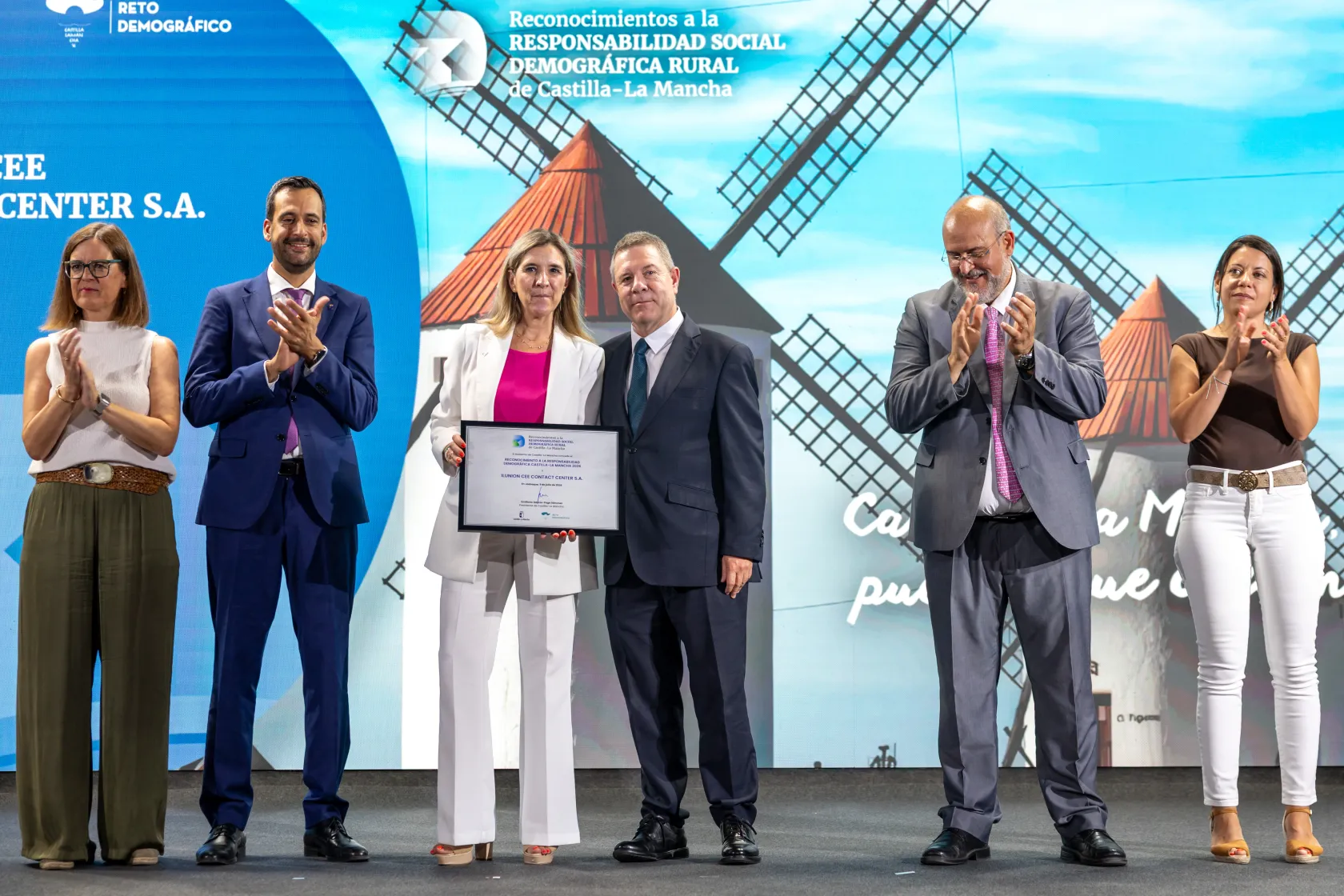 Entrega de reconocimiento a la responsabilidad social demográfica en Castilla-La Mancha; dos personas posan con diploma mientras otras aplauden frente a un escenario con molinos.