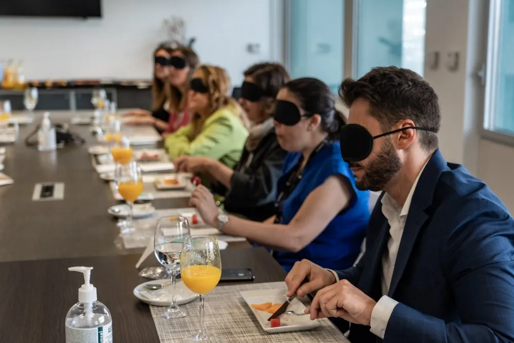 Varias personas participan en una experiencia de desayuno a ciegas alrededor de una mesa de reuniones. Todas llevan antifaces negros mientras degustan distintos alimentos y bebidas servidos en platos y copas. En primer plano, una persona sostiene cubiertos frente a un plato con fruta. La escena se desarrolla en una sala luminosa, con el objetivo de experimentar una situación similar a la de una persona con discapacidad visual.