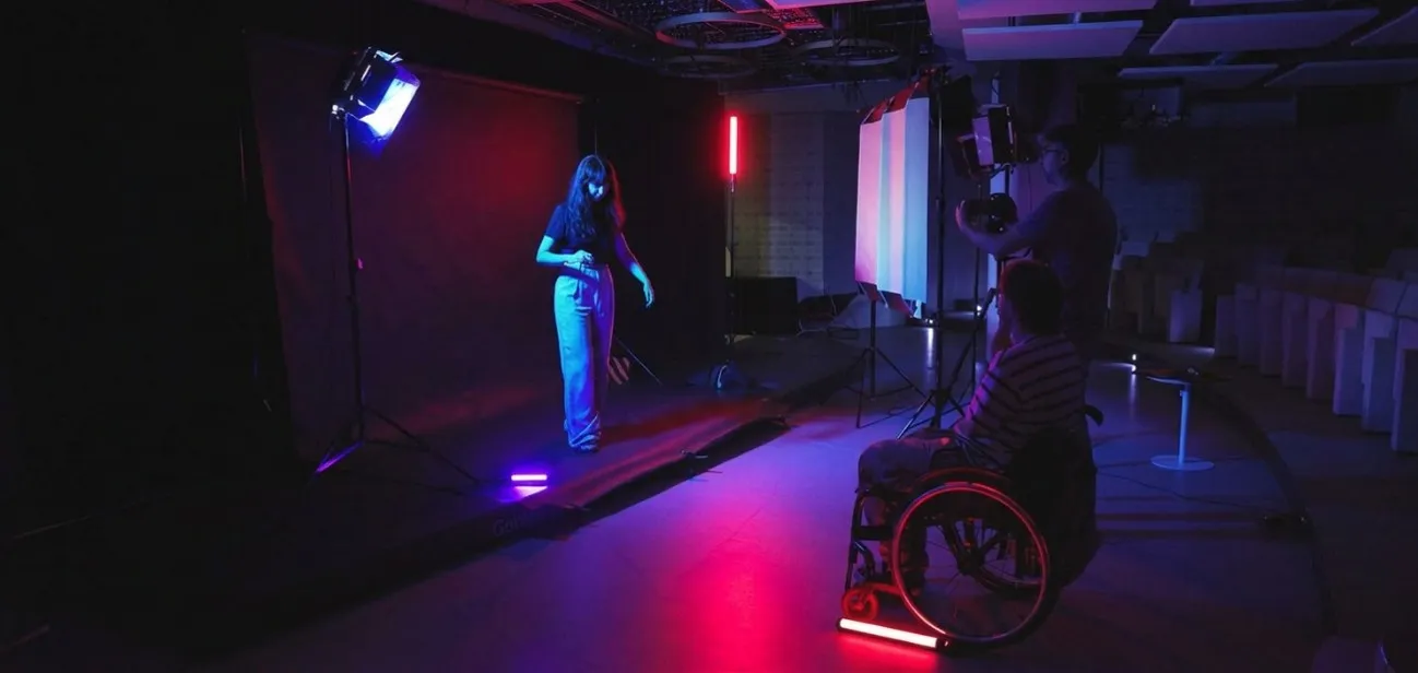 Grabación de un contenido audiovisual en un estudio, con una persona frente a cámara, equipo de producción y una persona usuaria de silla de ruedas observando la escena