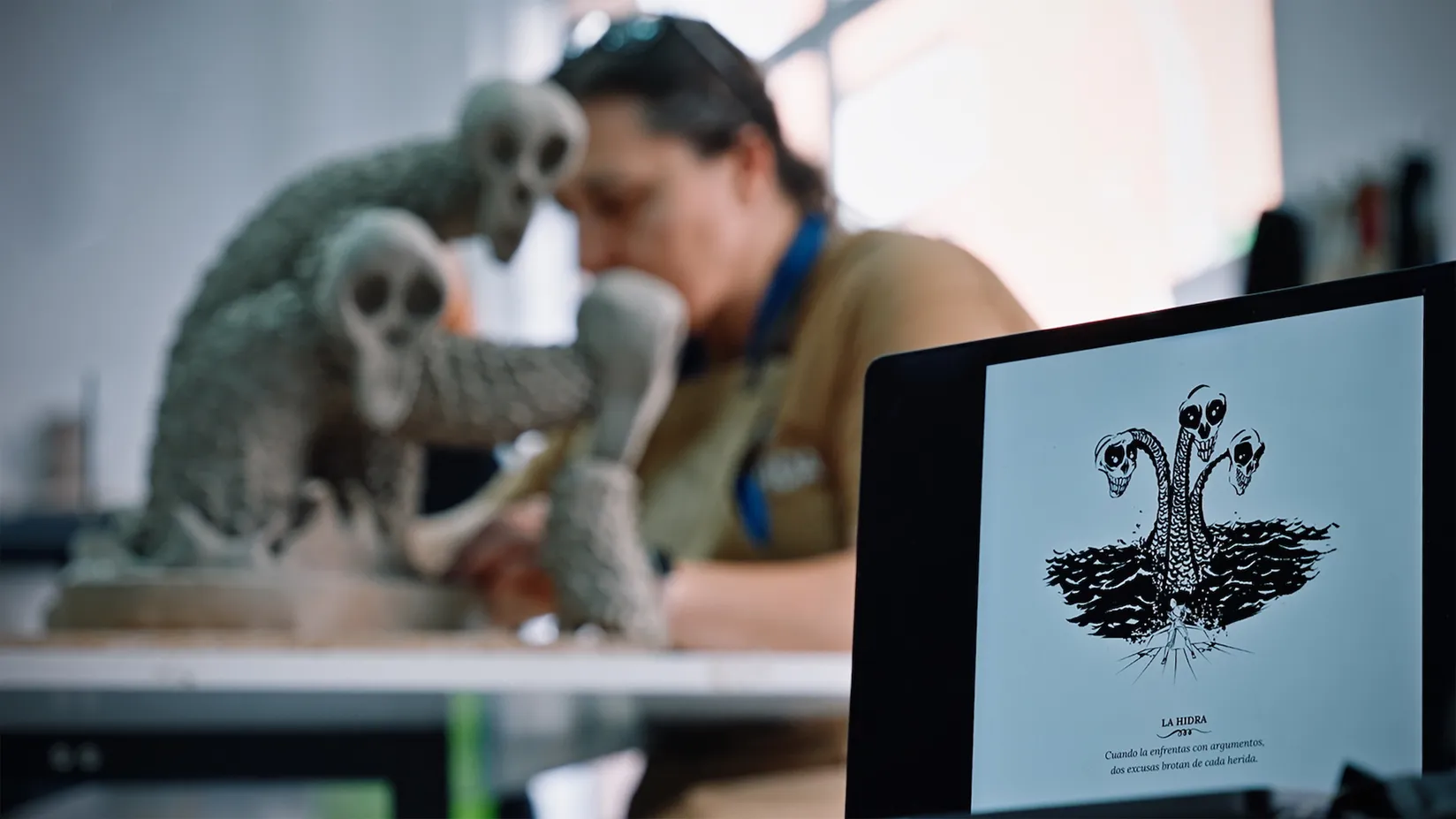 En primer plano, una tableta muestra el dibujo en blanco y negro de una hidra de varias cabezas. Al fondo, una persona trabaja en una escultura inspirada en esa misma criatura sobre una mesa de taller.