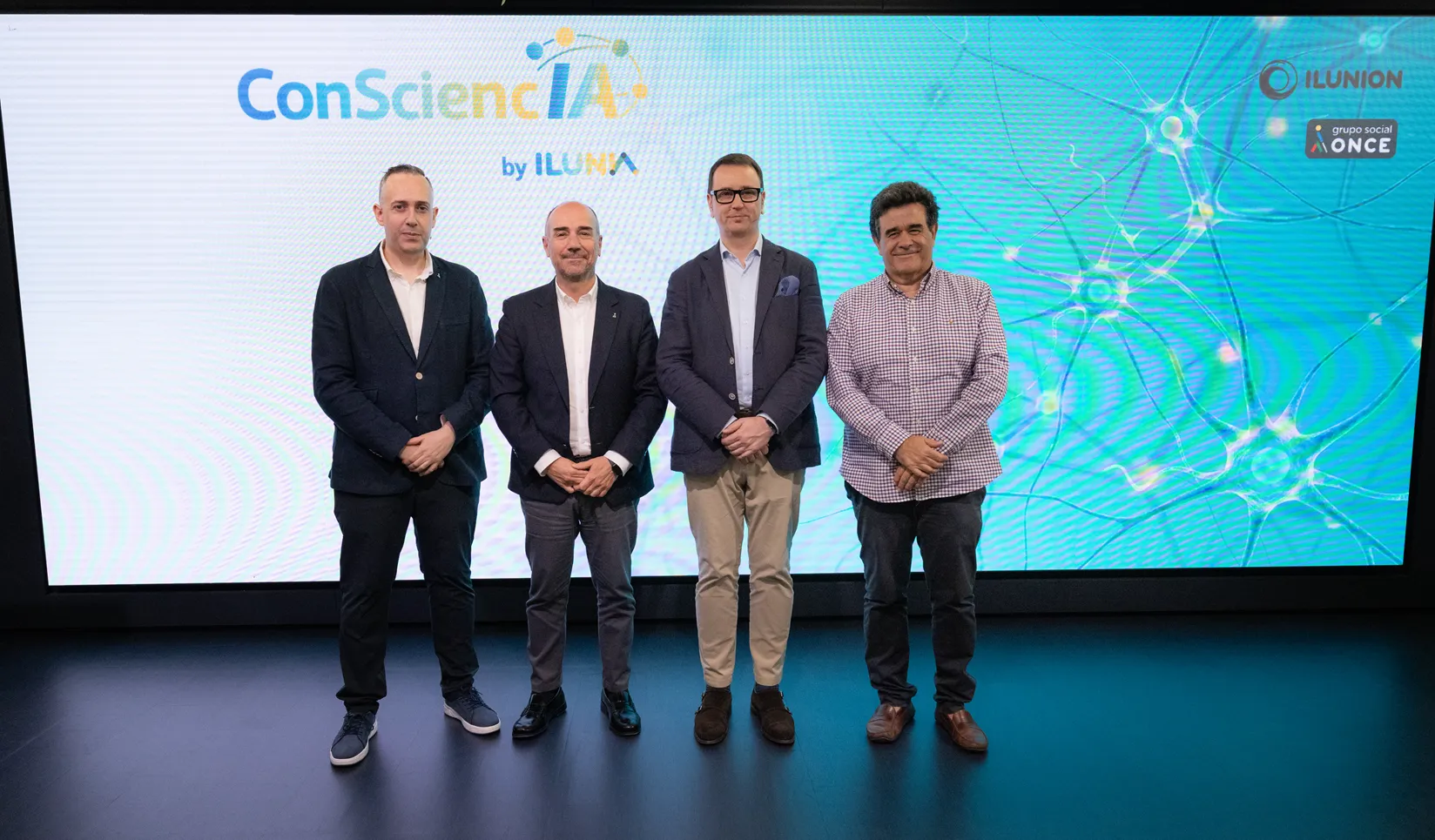 Cuatro personas posan de pie ante una gran pantalla durante una presentación de ConSciencIA by ILUNIA. En la pantalla, de tonos azul y verde, aparece una ilustración de conexiones neuronales y, en la esquina superior derecha, los logotipos de ILUNION y Grupo Social ONCE.