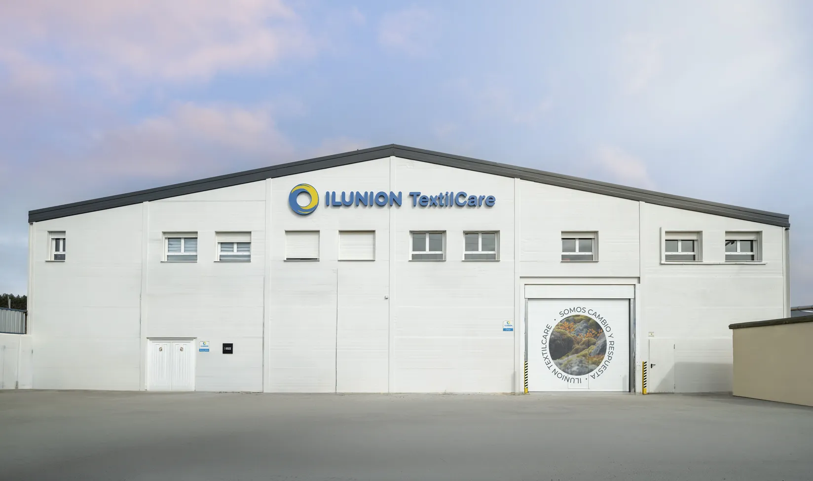 Exterior de una planta de ILUNION TextilCare. El edificio, de fachada blanca, muestra el logotipo de la empresa en la parte superior y varias ventanas. A la derecha hay una gran puerta de acceso con una imagen de paisaje y el mensaje “Somos cambio y respuesta”.