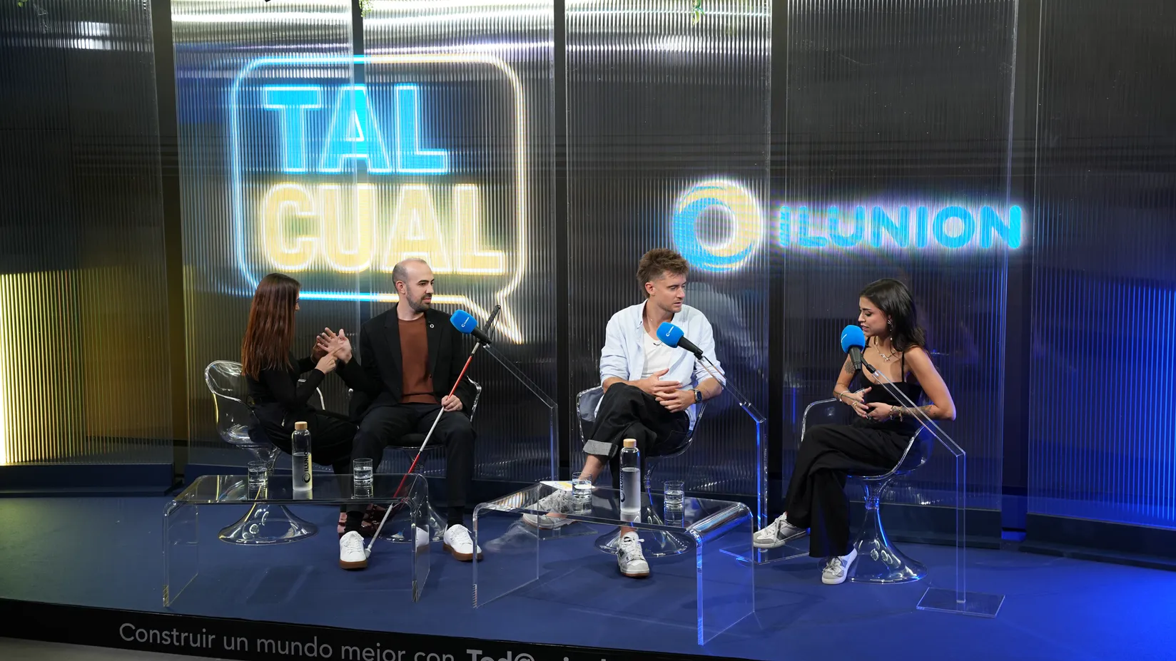 Cuatro personas conversan sentadas en un escenario, ante varios micrófonos. Una de ellas sostiene un bastón blanco y rojo mientras interactúa con la persona sentada a su lado. Al fondo se ven iluminados los textos «TAL CUAL» e «ILUNION».