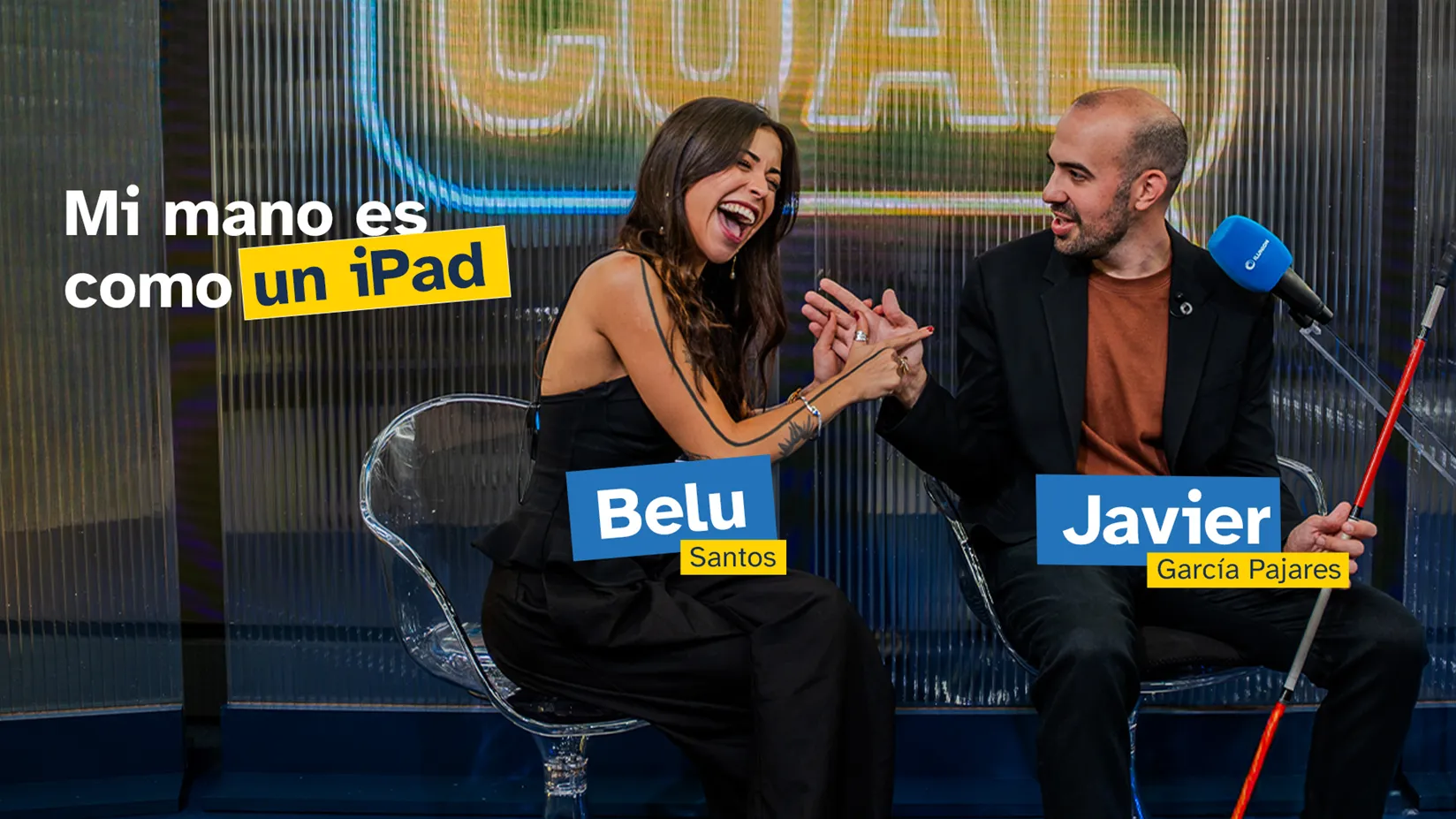 Dos personas sentadas conversan ante un micrófono. Una de ellas toca con las manos las de la otra, que sostiene un bastón blanco y rojo. Sobre la imagen aparece el texto «Mi mano es como un iPad» y los nombres «Belu Santos» y «Javier García Pajares».