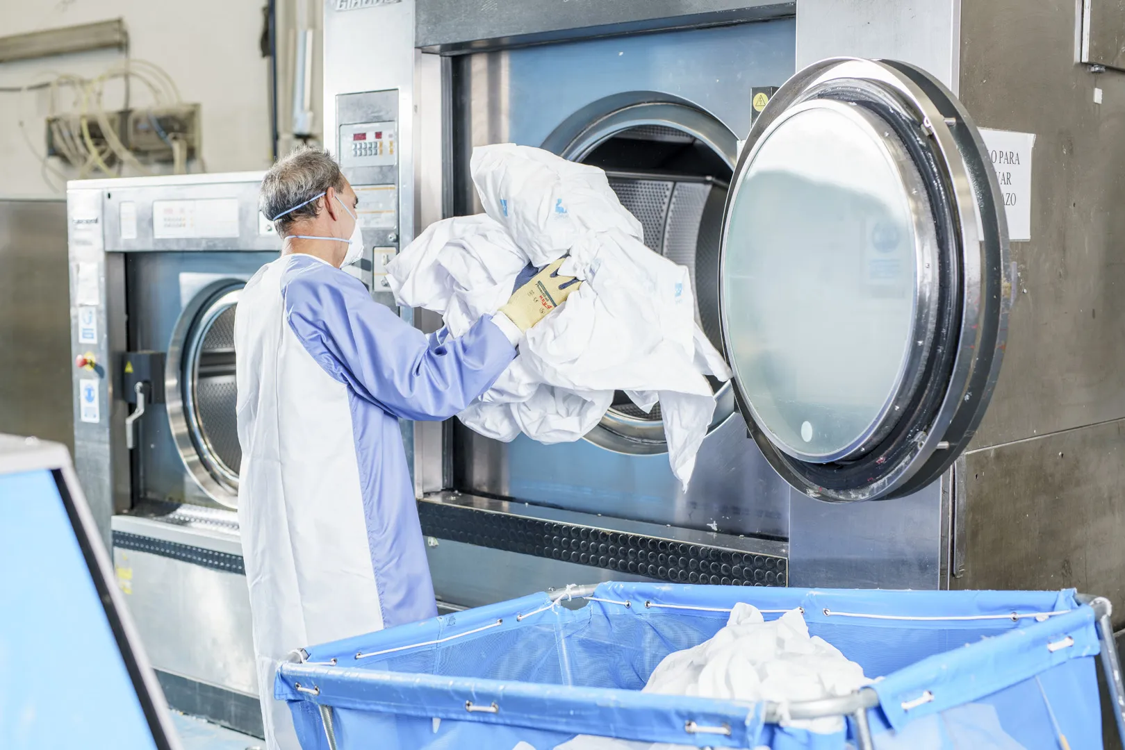 Imagen de un trabajador con su EPI introduciendo ropa de cama en una lavadora 
