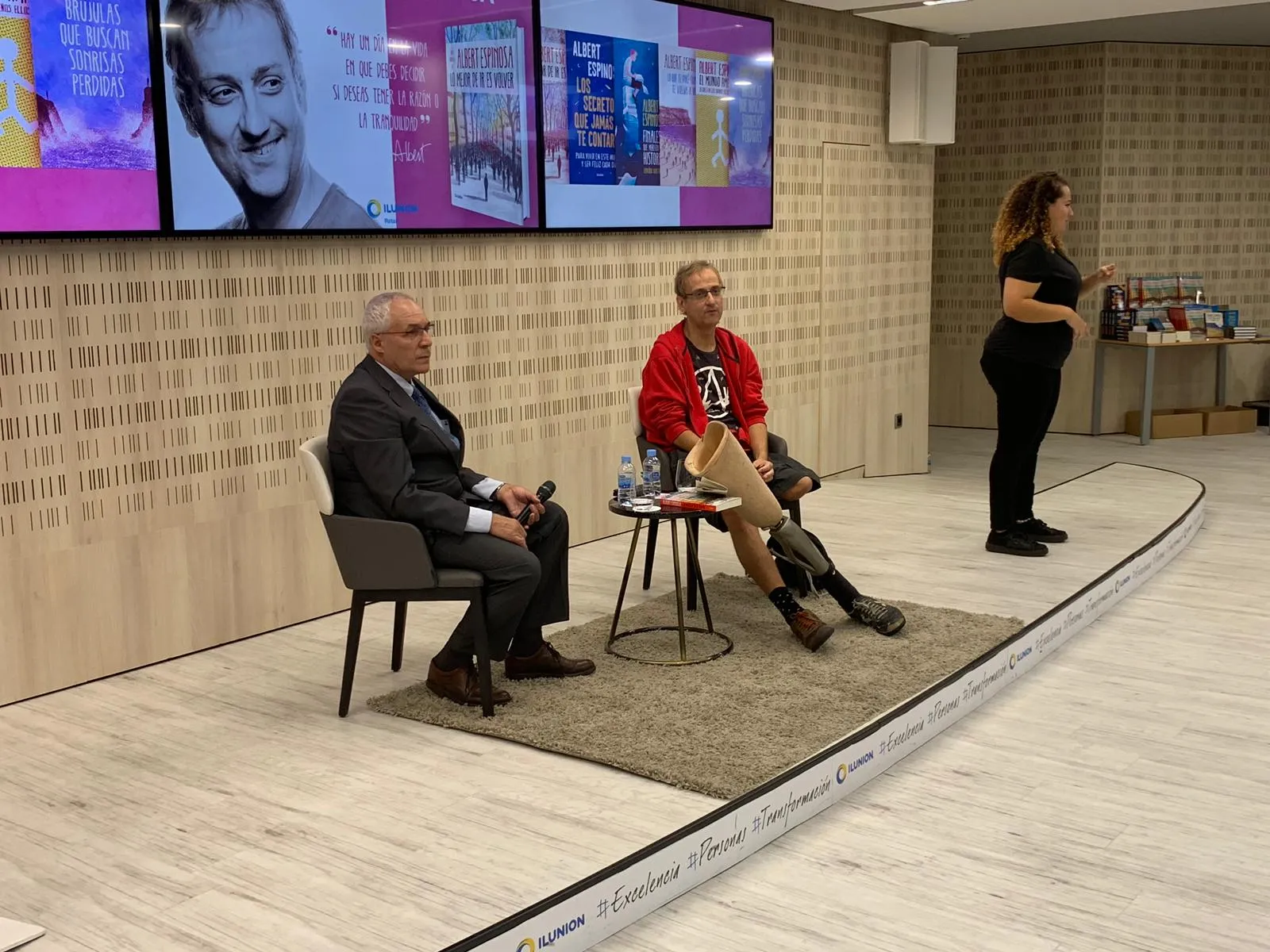Albert Espinosa en el salón de actos de Torre ILUNION. Él está sentado, con su pierna ortopédica apoyada en la mesa. A un lado, una intérprete de lengua de signos. Está acompañado en el escenario por una persona de ILUNION, que está vestida con traje de chaqueta y corbata y que también está sentada. Cada uno en una silla. 