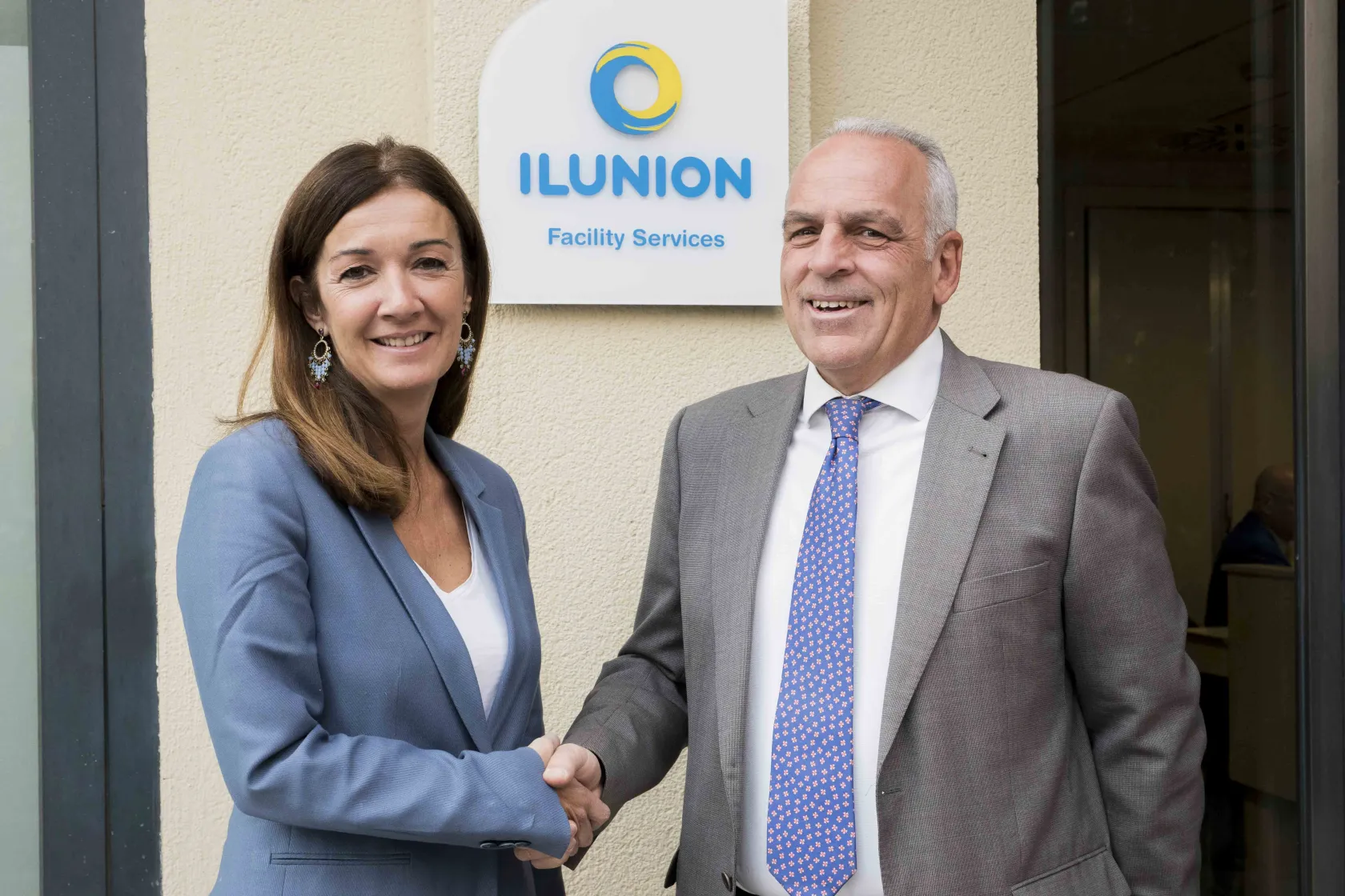 Imagen en donde se observa a la directora de APRAMP, Rocio Mora, y al director de Recursos Humanos de ILUNION Facility Services, Pedro Buesa, en la entrada de la delegación de ILUNION Facility Services en Madrid
